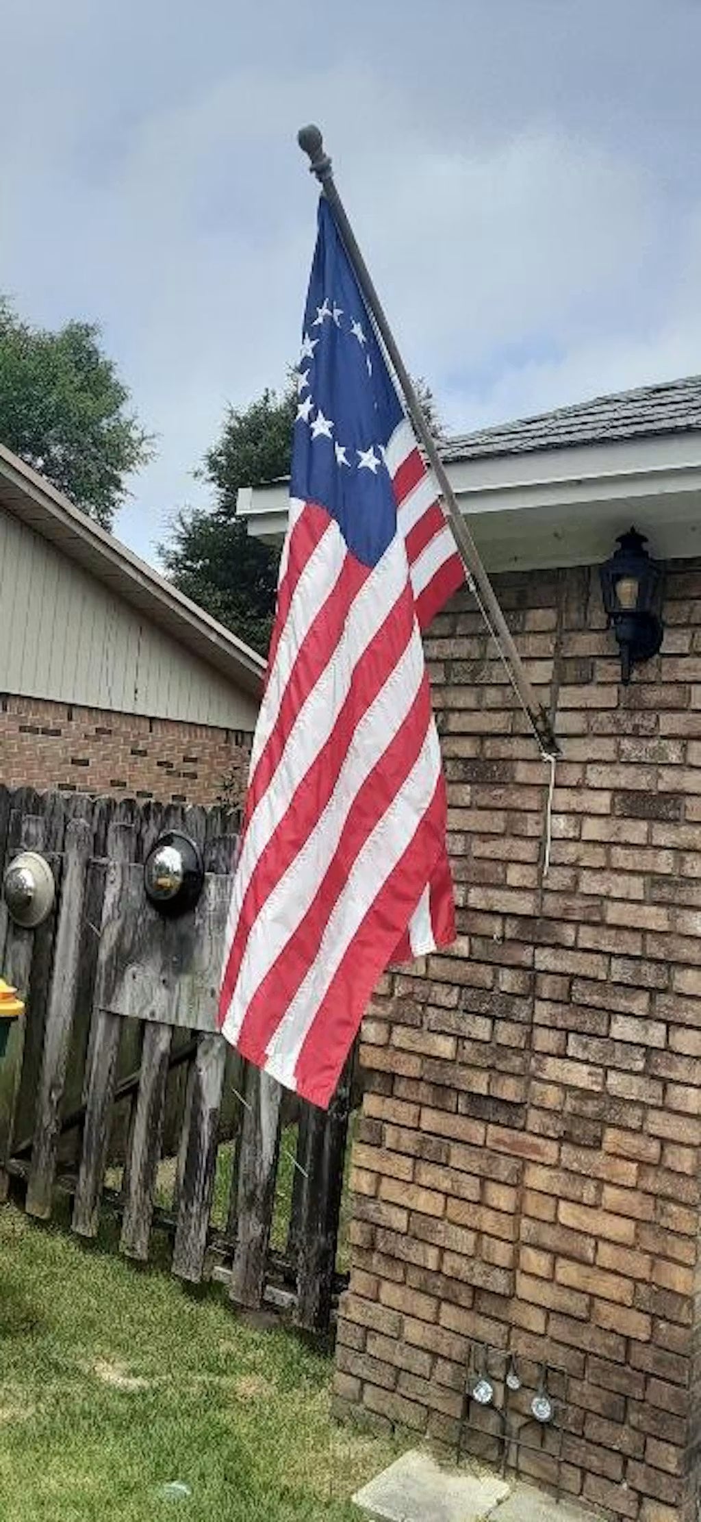 Besty ross flag on a flagpole in front of a brick building with a wooden fence.