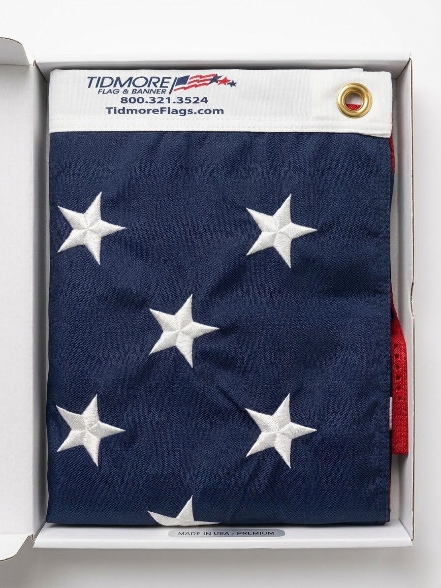 Folded American flag with white stars on a blue background, displayed in a box with Tidmore Flags branding.
