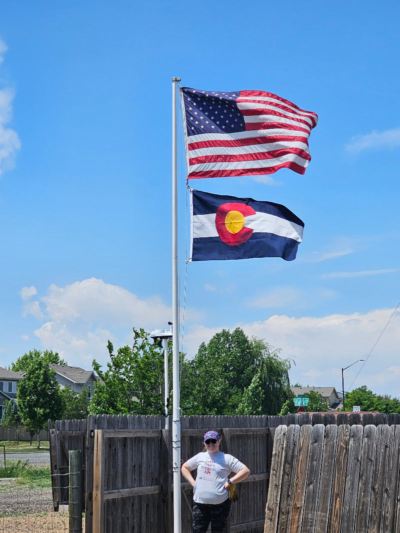 Two flags, American and Colorado, waving against a blue sky with a wooden structure and person in the background.